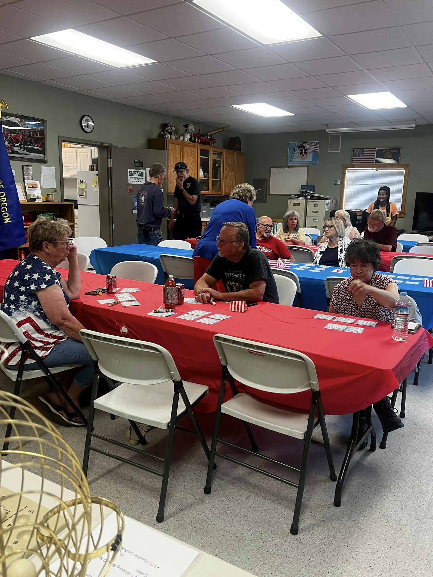 People sitting at tables with red and blue covers, likely in a community center or gathering, some are drinking from bottles.