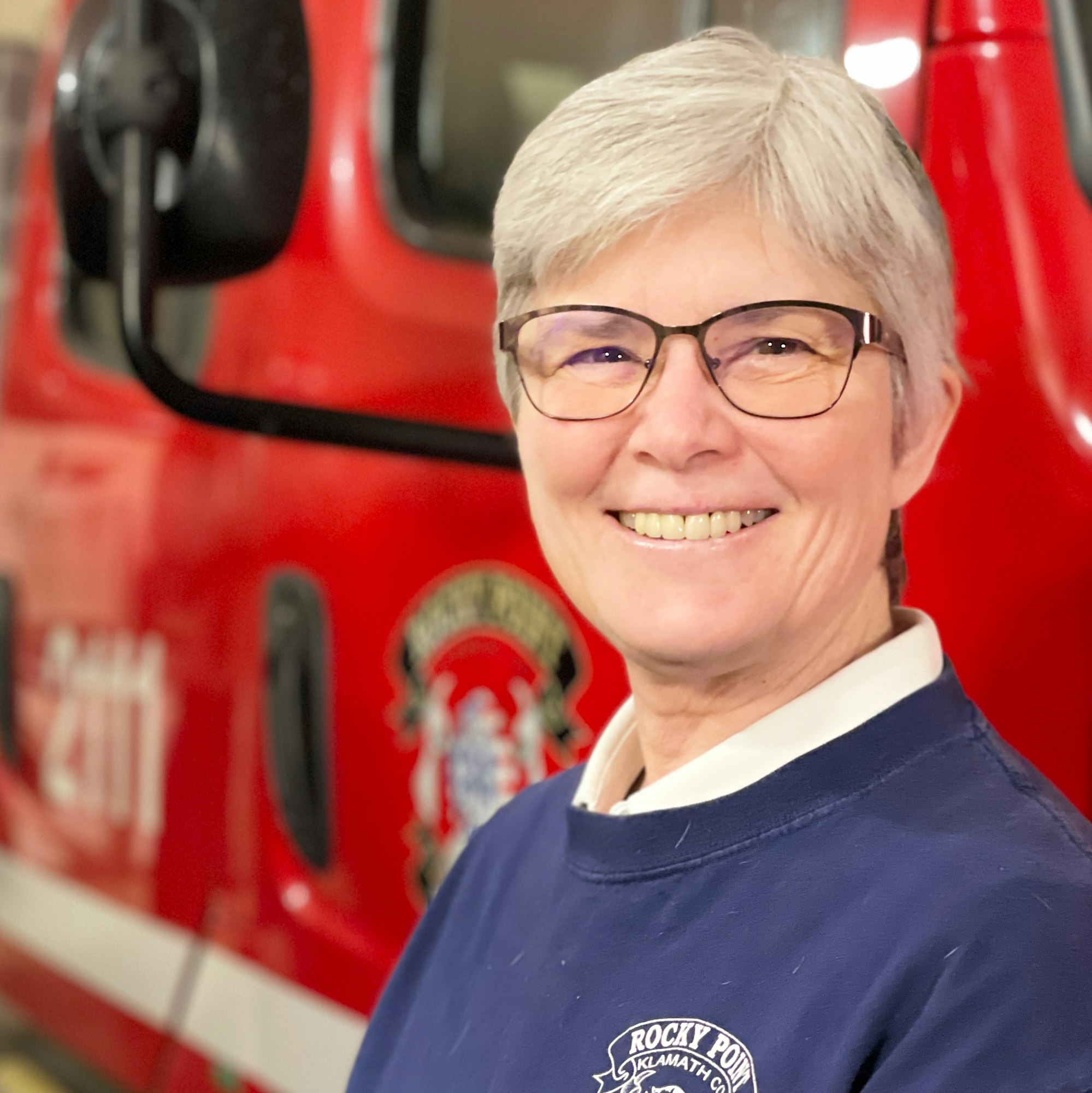 A smiling person with short gray hair and glasses stands in front of a red fire truck, wearing a dark blue sweatshirt.