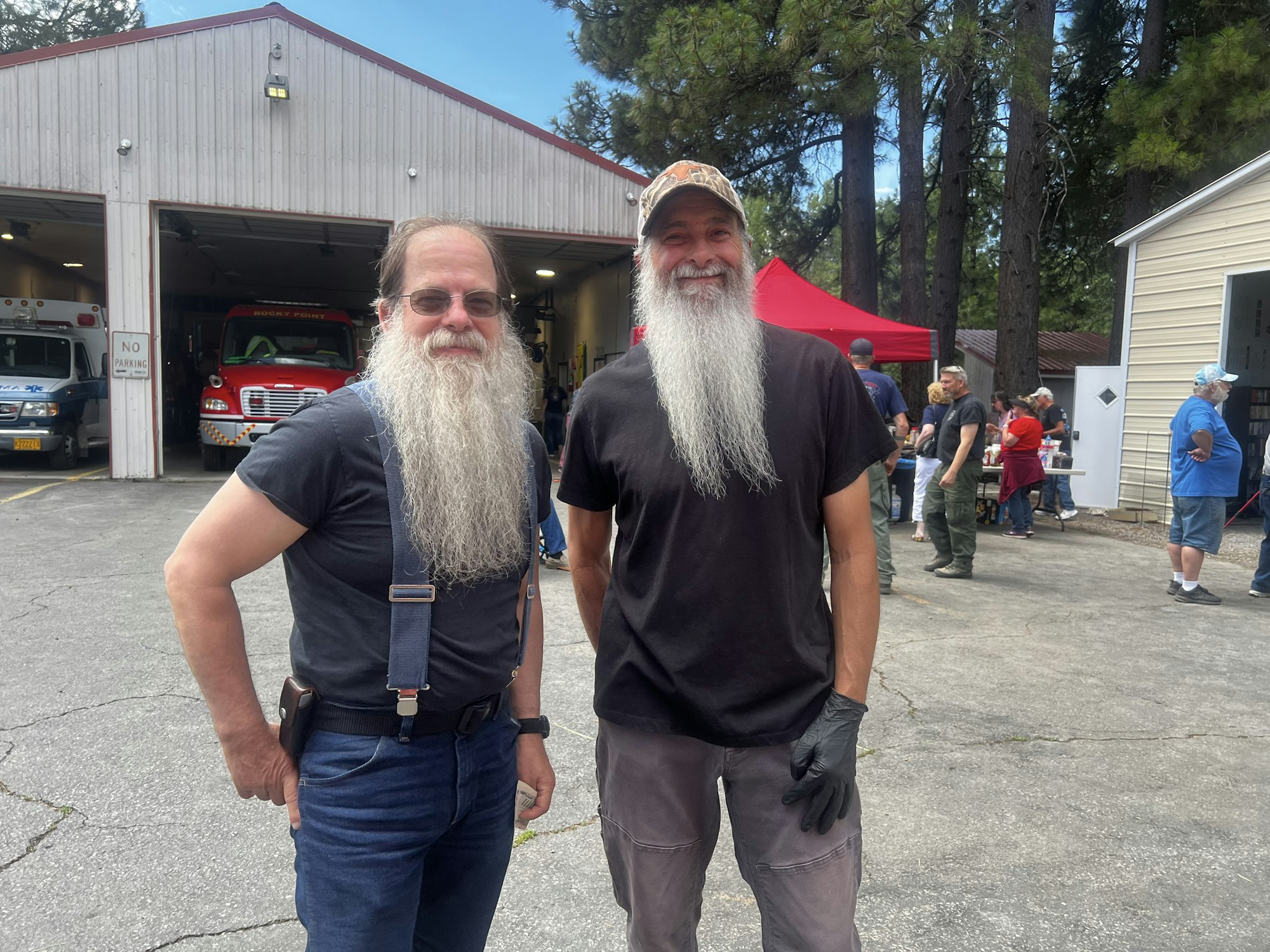 Two men with long beards stand smiling outside a garage with emergency vehicles, surrounded by people and trees.