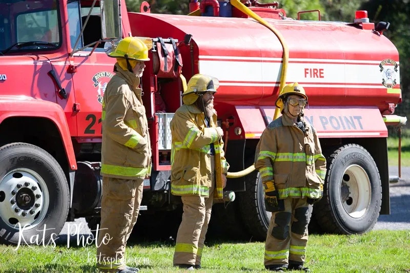 Three firefighters in gear stand beside a red fire truck labeled "FIRE."