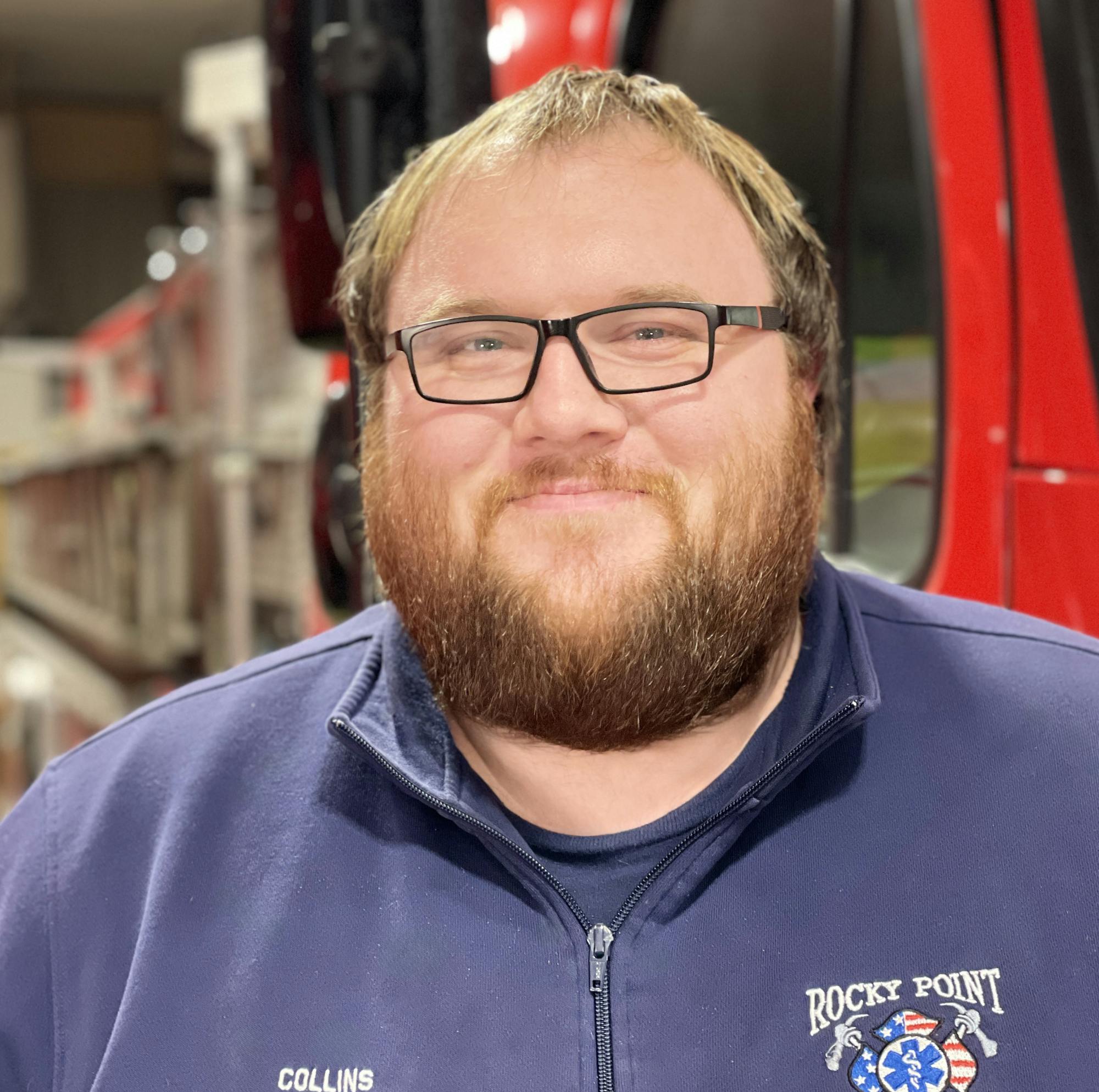 A bearded man in a navy jacket with "Rocky Point Fire EMS" and "COLLINS" stands in front of a fire truck.