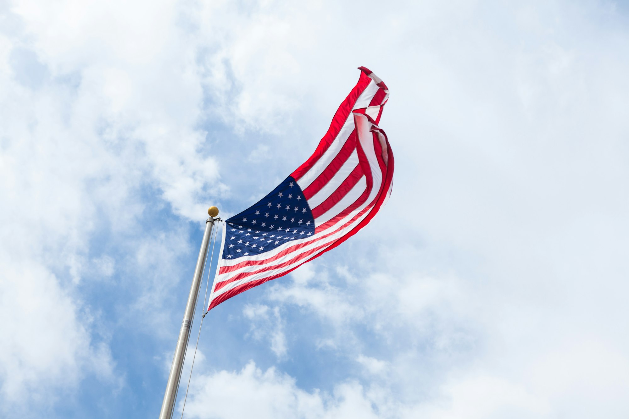 A U.S. flag waving on a flagpole against a cloudy blue sky.