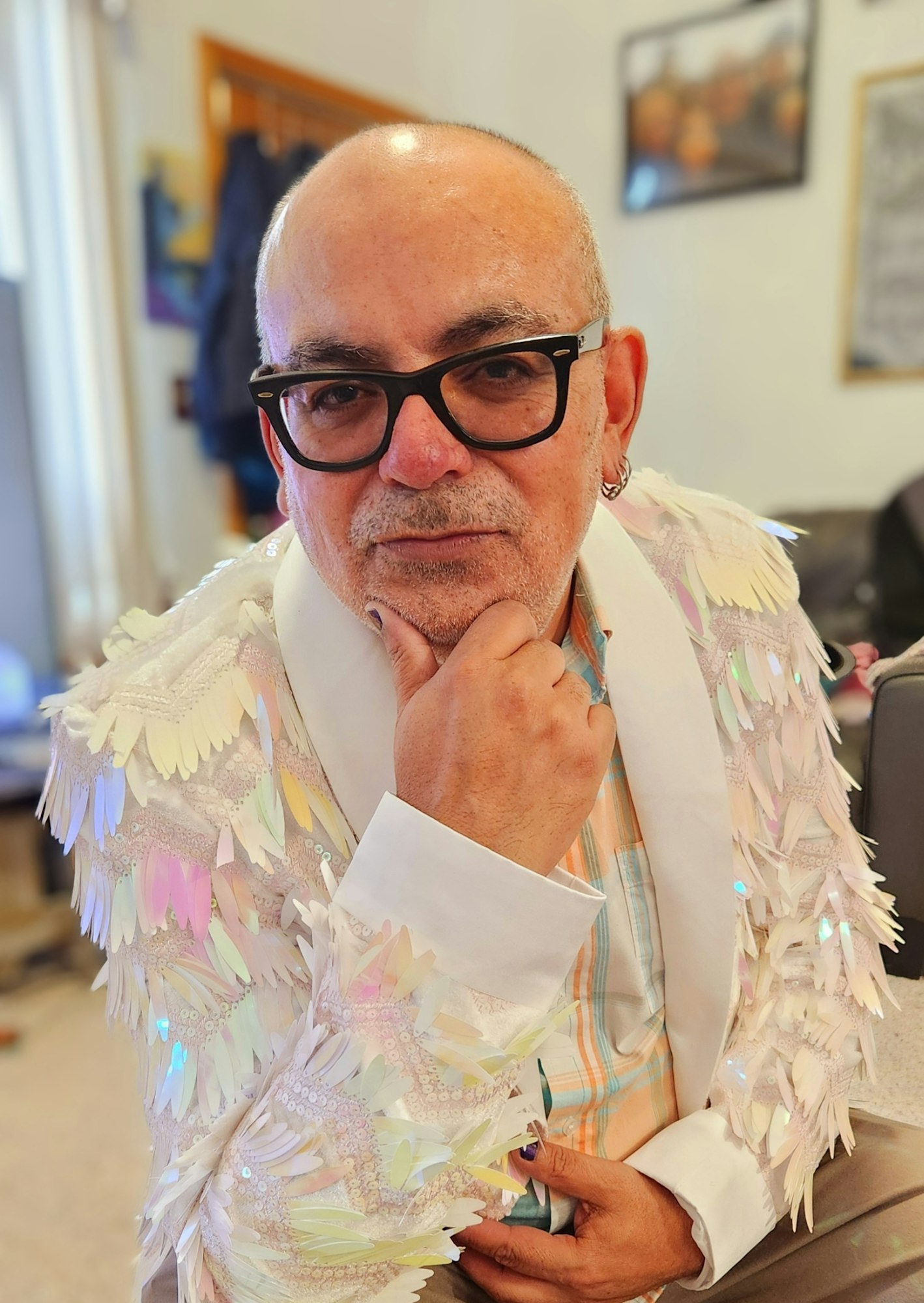 A person wearing glasses and a colorful sequin jacket, striking a thoughtful pose indoors.