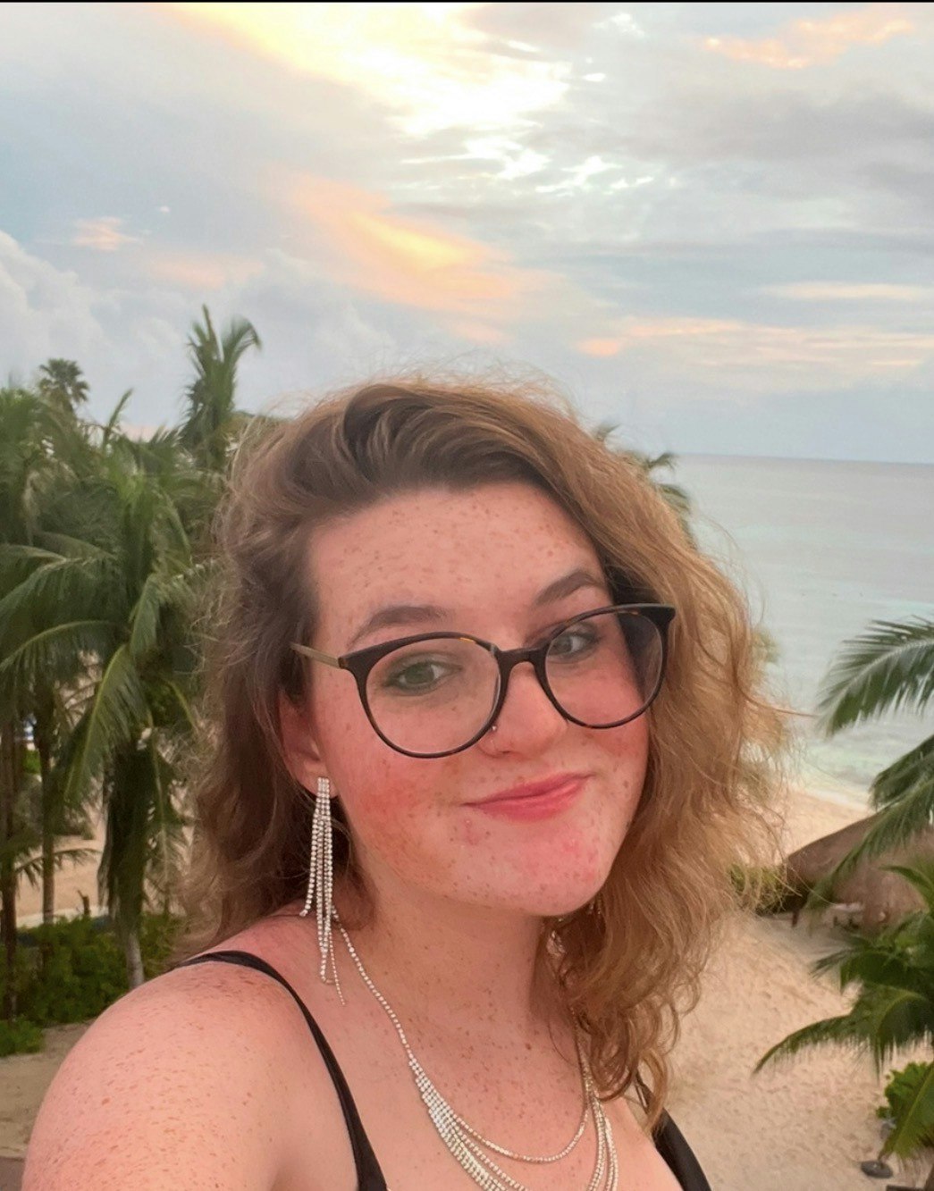 A person with curly hair and glasses smiles in front of a beach with palm trees and a colorful sky at sunset.