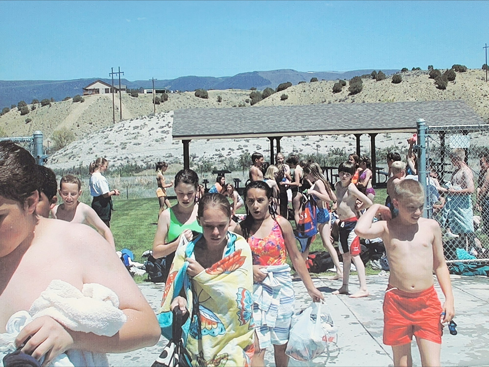 People at a poolside area, likely after swimming, some wrapped in towels, with hills in the background.
