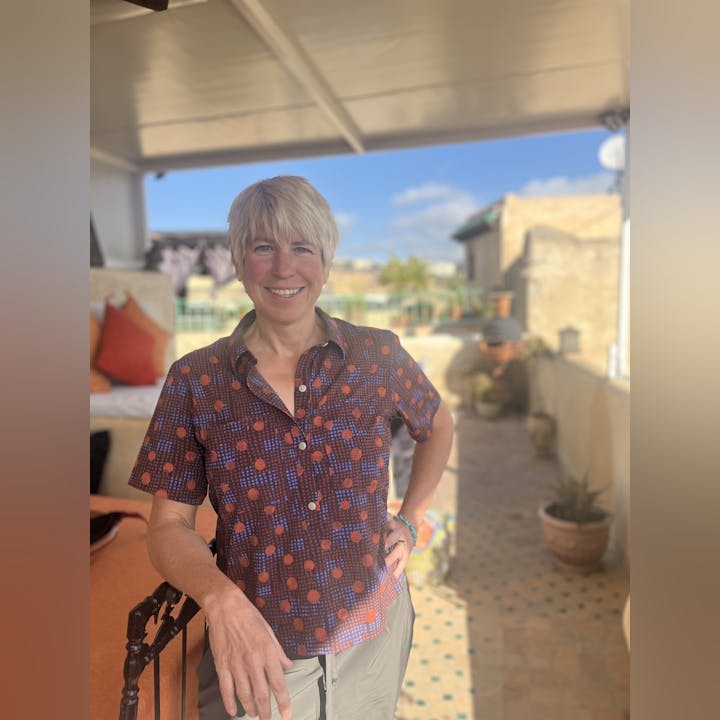 A smiling person standing on a sunny patio with plants and outdoor furniture.