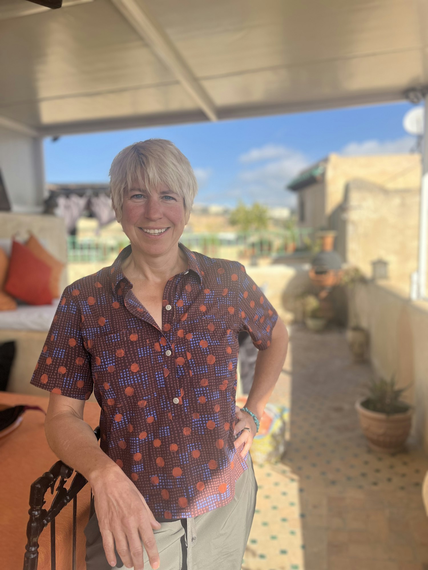 A smiling person standing on a sunny patio with plants and outdoor furniture.
