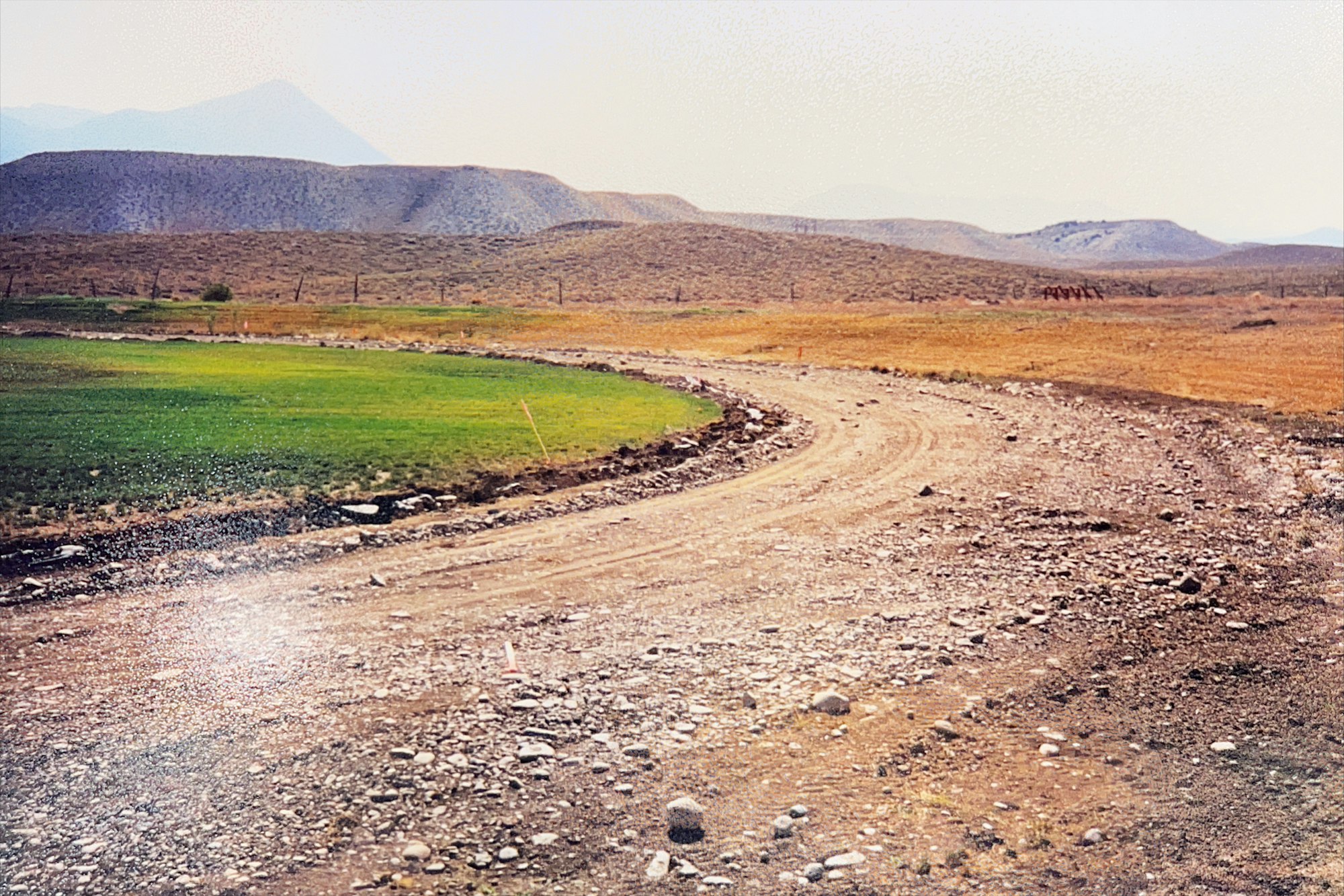 A curving dirt road through a dry landscape with hills and a patch of greenery.