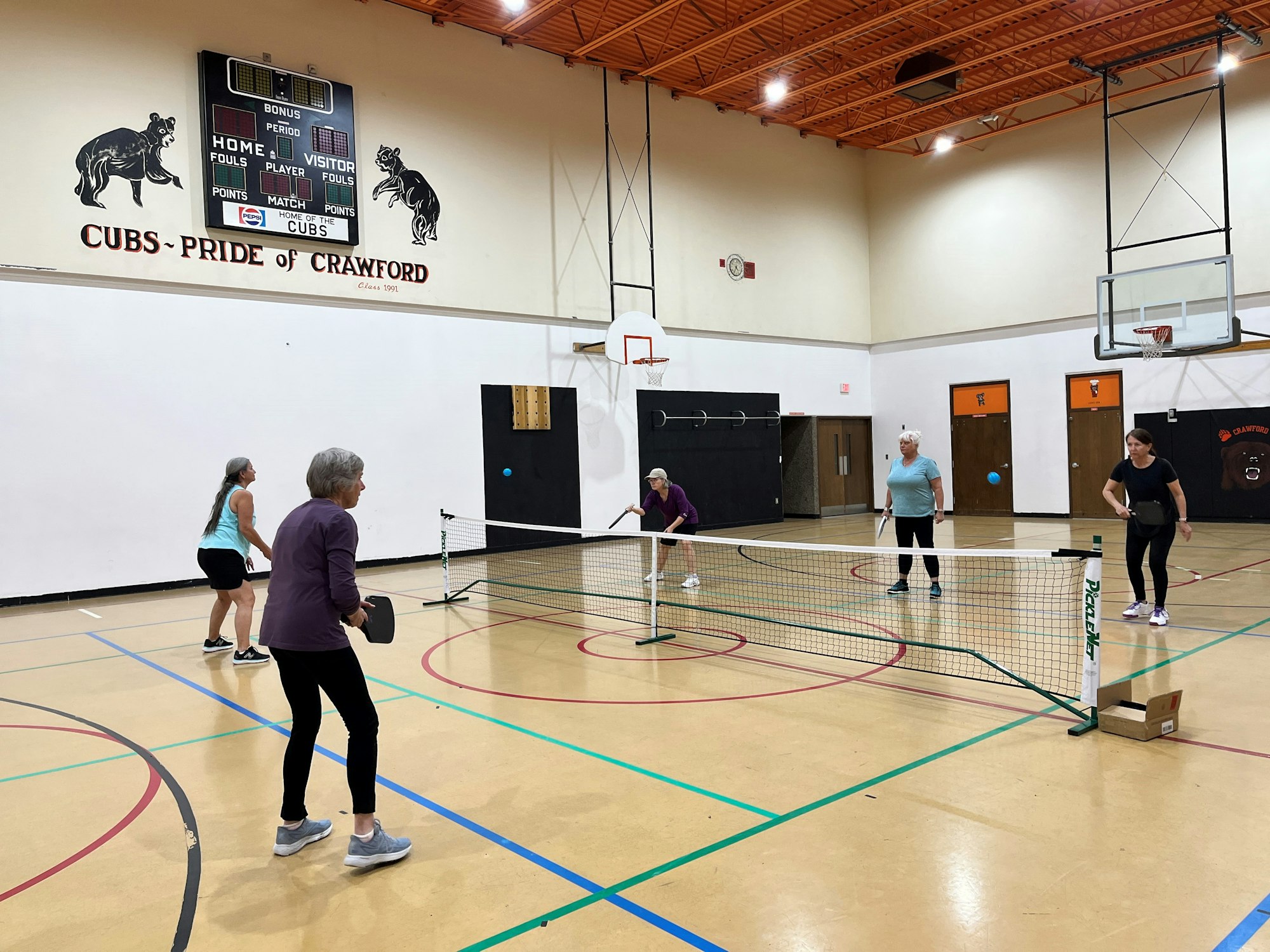 People playing pickleball in a gym with a scoreboard showing "CUBS - PRIDE OF CRAWFORD."