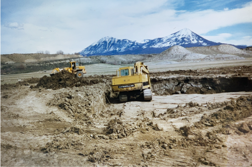 Construction site with two bulldozers, excavated land in the foreground, snow-capped mountains in the background.