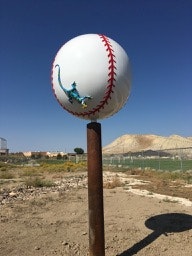 A large baseball sculpture with a dinosaur toy on top, set against a landscape with mountains in the background.