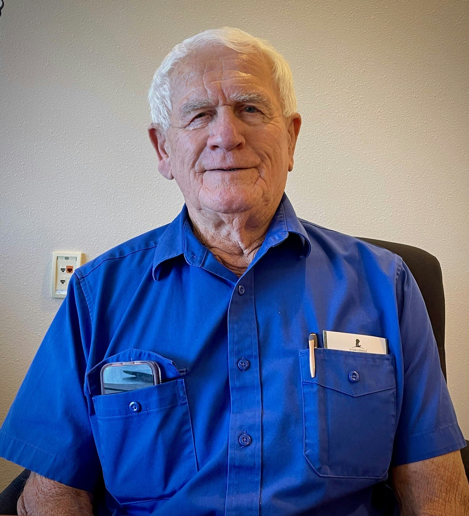 A gentelman in a blue shirt sits against a neutral background, with a phone and pen in his shirt pockets.