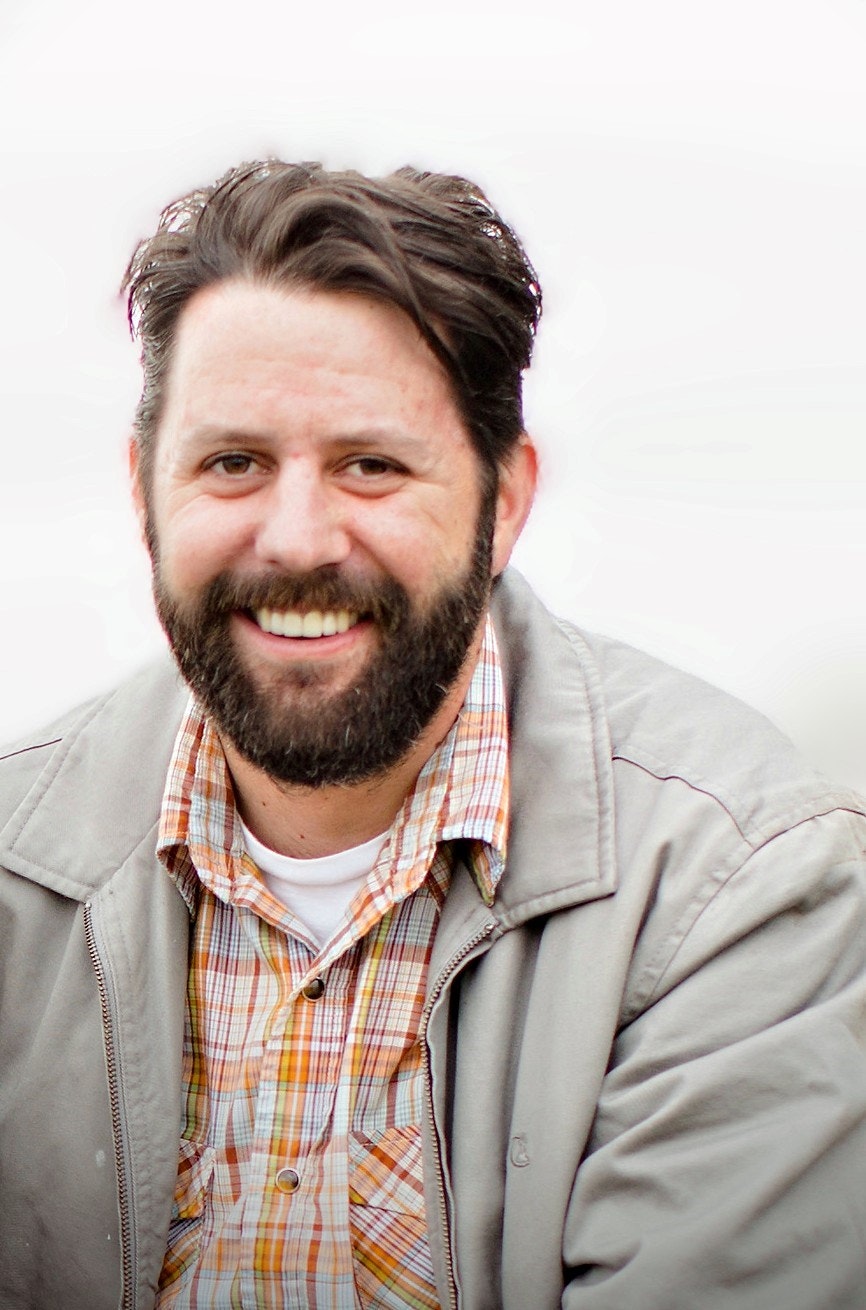 A smiling man with a beard in a plaid shirt and jacket.