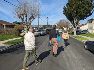 Three people walking down a sunny street lined with houses, trees, and parked cars.
