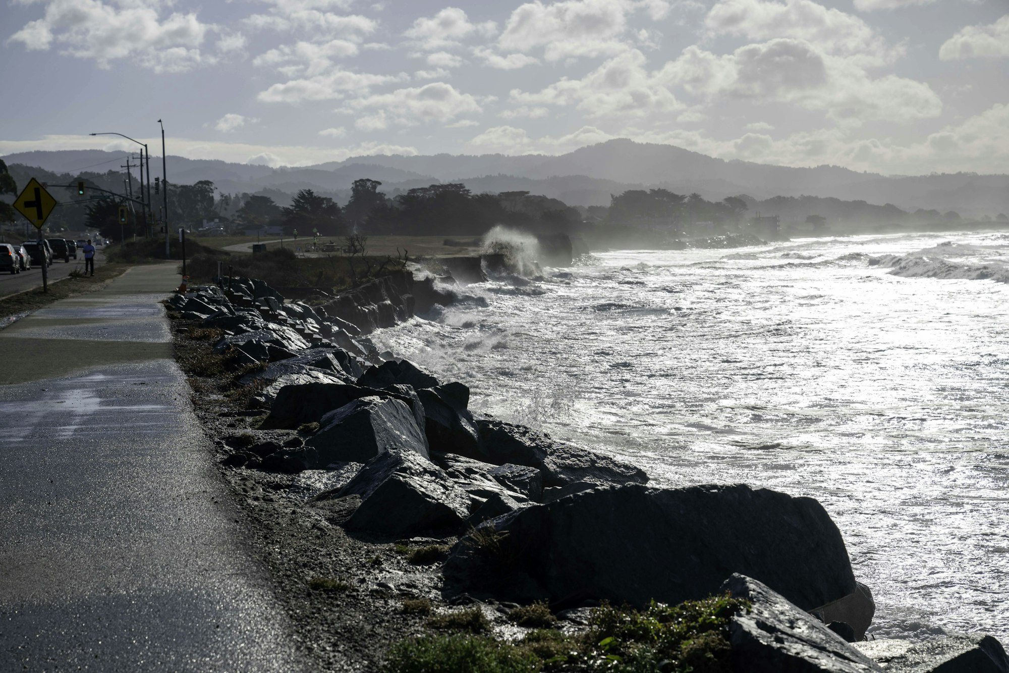 The image features a rocky shoreline with waves crashing against it, a path beside the ocean, and mountains in the background.