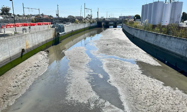The image shows a shallow, muddy waterway with industrial structures and train tracks in the background.