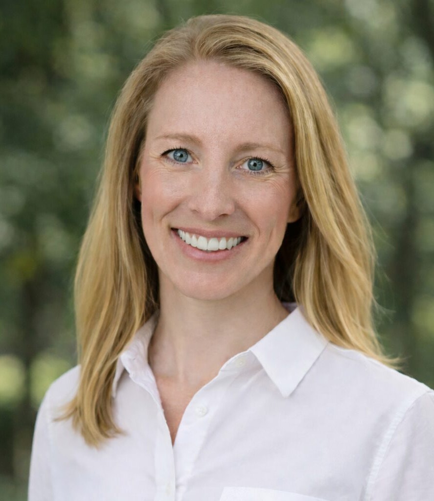 The image features a smiling woman with long, light brown hair, wearing a white shirt, set against a blurred green background.