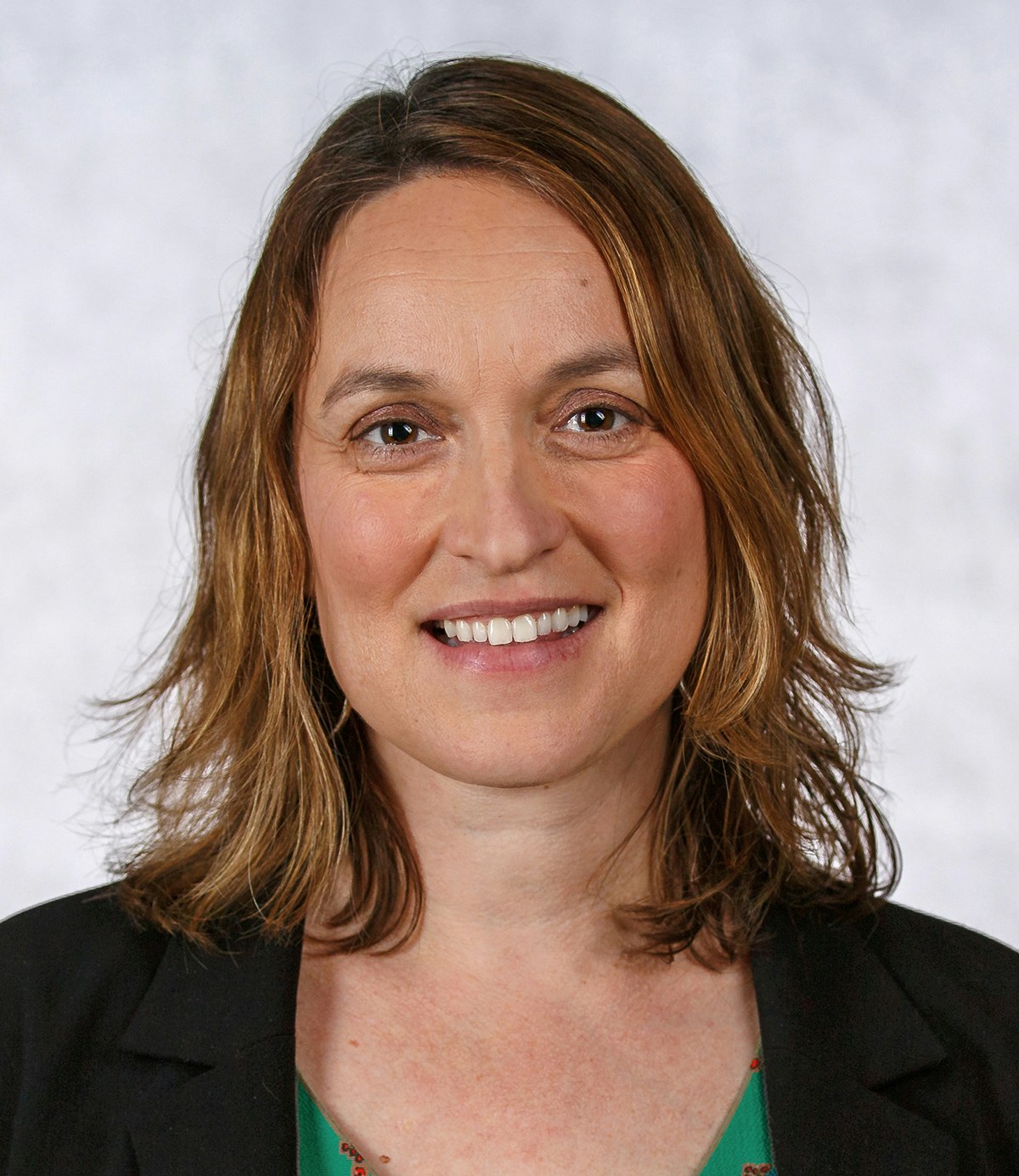 The image features a woman with medium-length wavy hair, smiling, wearing a green top and a black blazer, set against a light background.