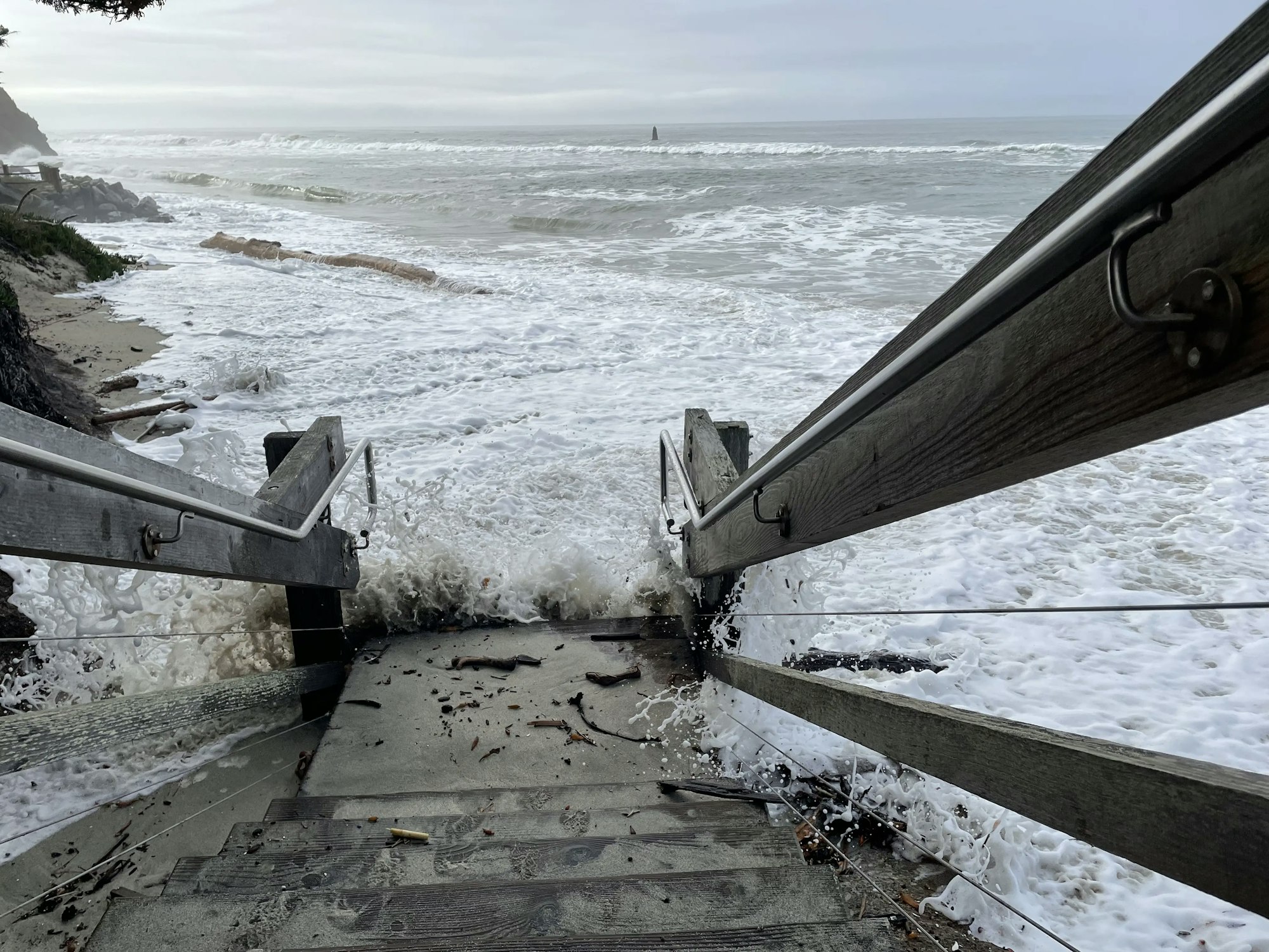 A set of wooden stairs leads down to a beach, with waves crashing at the bottom, creating foam and debris on the steps.