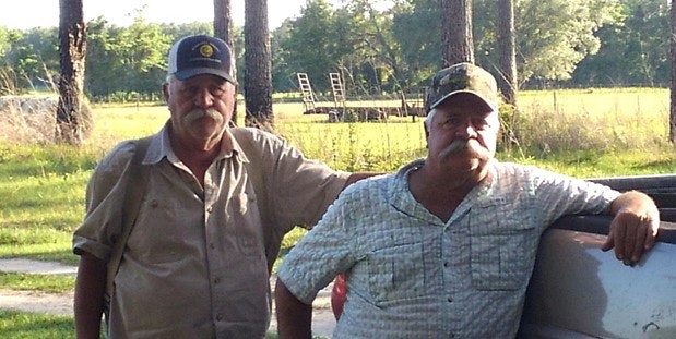 Two men with mustaches wearing caps stand outdoors near a vehicle, with trees and grass in the background.