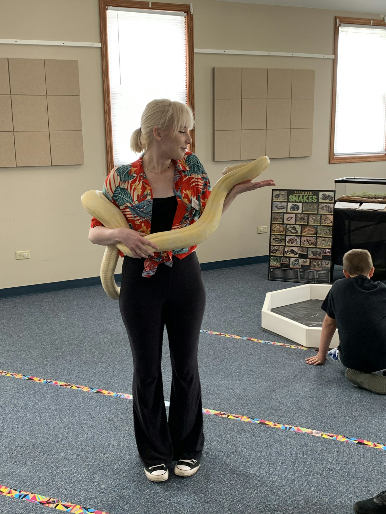 A person holding a large yellow snake in a room; another person is kneeling on the floor.