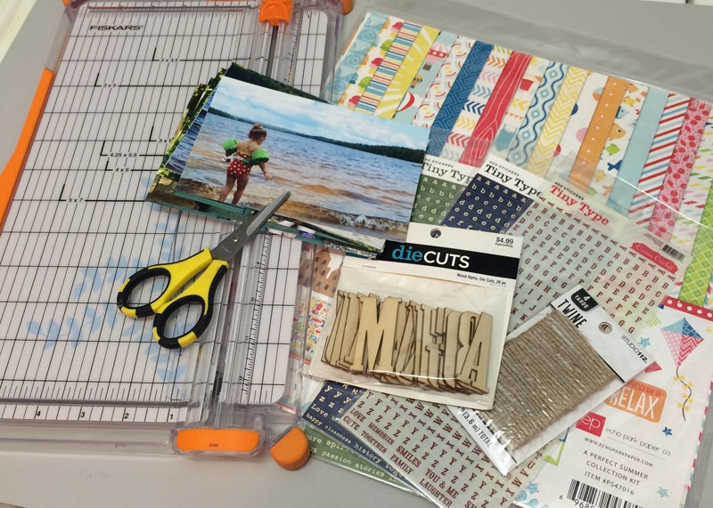 Scrapbooking supplies: paper trimmer, scissors, photos, patterned paper, die cuts, twine, and letter stickers.