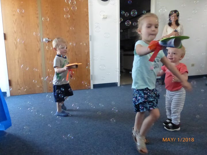 Children are playing in a room filled with bubbles, with one girl holding a colorful bubble wand and two boys watching happily.