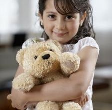 A young girl is smiling while holding a teddy bear close to her chest. The setting appears warm and inviting.