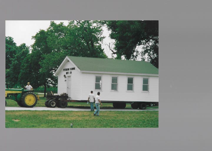 A white schoolhouse on a trailer being moved, with a tractor nearby and people overseeing the process.