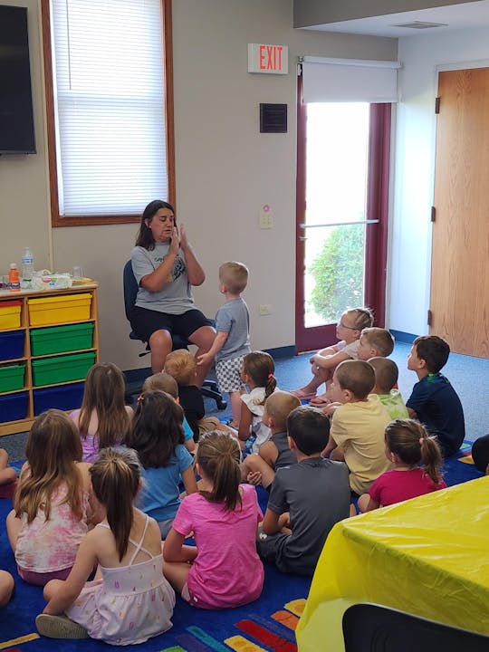 Children sitting on the floor, focused on a woman who might be teaching or storytelling. The room looks like a classroom.