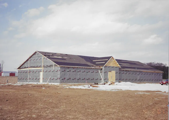 The image shows a partially constructed building with a gray exterior and an unfinished roof, set in a rural area.
