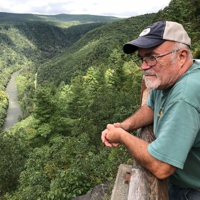 A man in a cap and glasses overlooks a lush, forested valley with a winding river from a wooden railing.