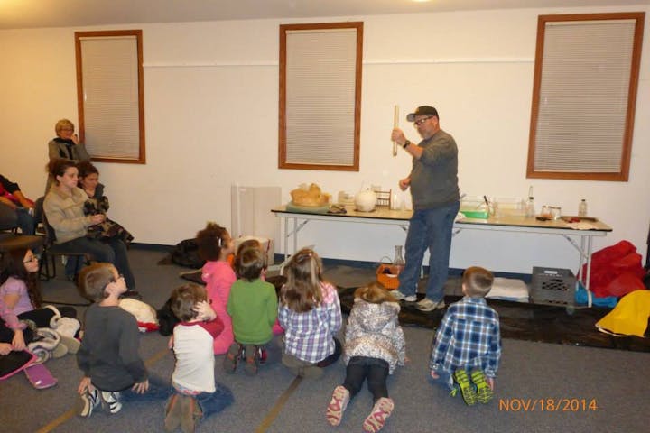 A man conducts a demonstration for children seated on the floor, while adults observe in the background.