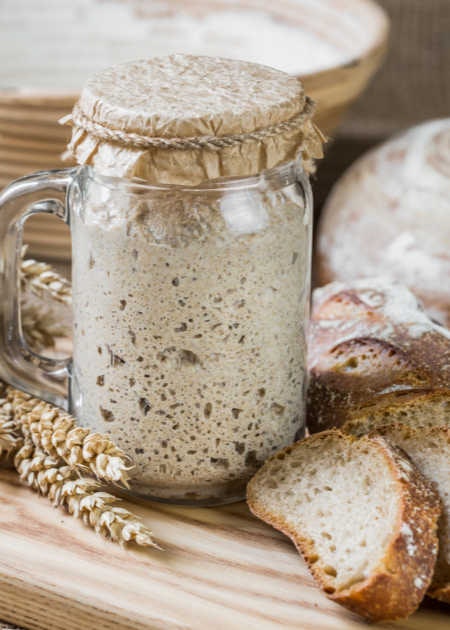 A jar of sourdough starter next to slices of bread and wheat, placed on a wooden surface.