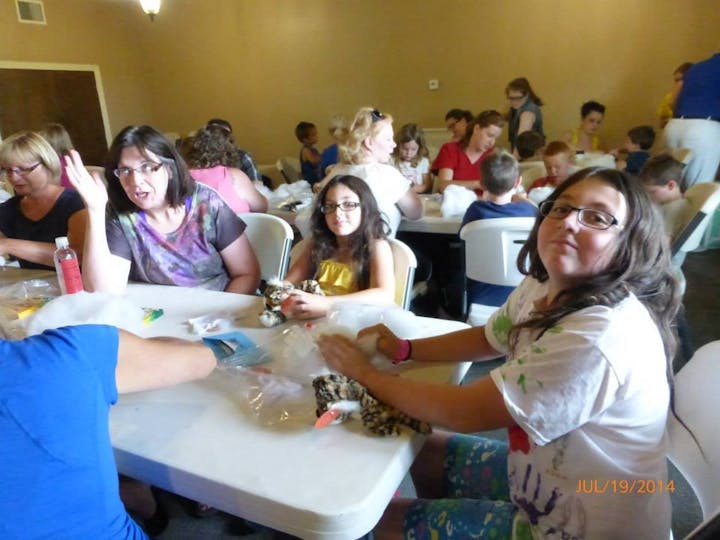 The image shows a group of people, including children and adults, engaged in an activity at tables, likely crafting or making something.