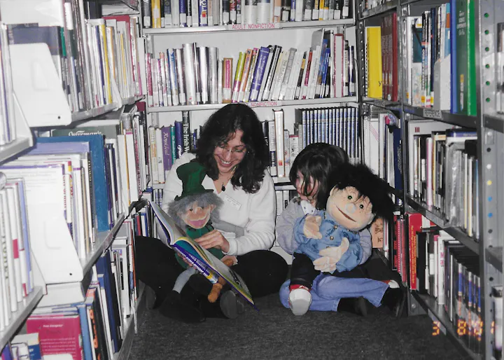A woman and a child sit among books in a library, reading a book with puppets, creating a fun storytelling atmosphere.