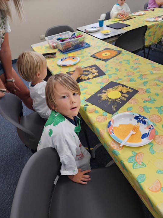 Child at a table with art supplies and colorful paintings; the image is rotated.