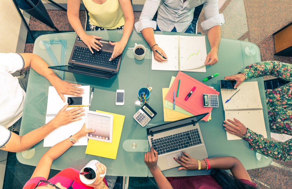 People working at a table with laptops, notebooks, calculators, and coffee.
