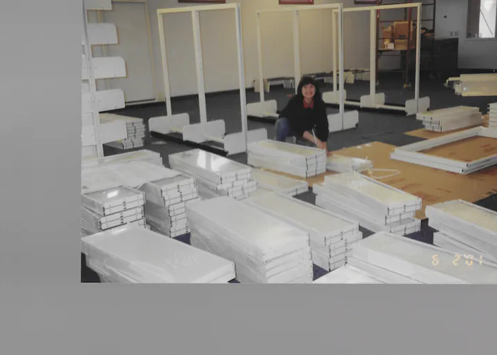 The image shows a woman assembling display furniture in a room filled with neatly stacked white shelves and parts.
