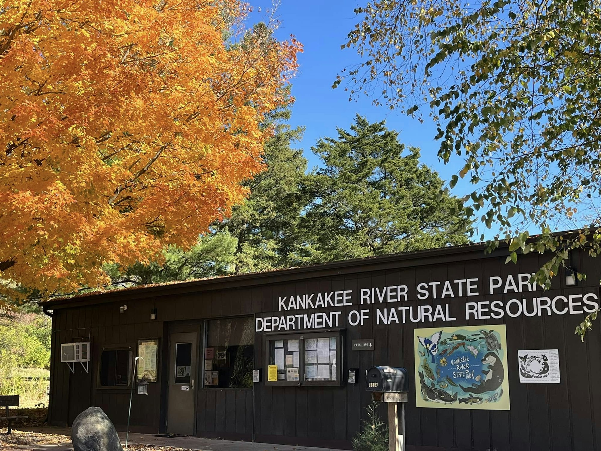 A building marked "KANKAKEE RIVER STATE PARK" with vibrant autumn trees and a clear blue sky.