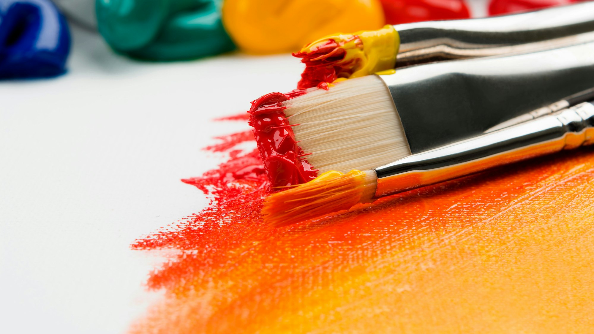 The image features paintbrushes with vibrant red and orange paint on a canvas, surrounded by colorful paint tubes.