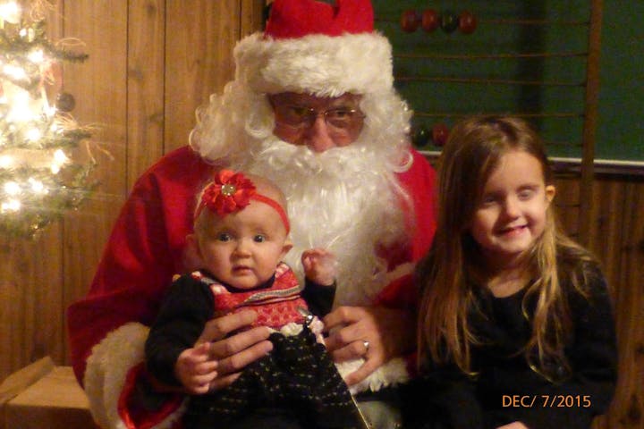 A man dressed as Santa holds a baby and a girl, smiling in front of a Christmas tree, capturing a festive moment.