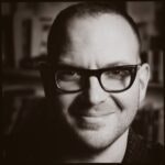 A person wearing glasses, smiling, in a sepia-toned portrait.