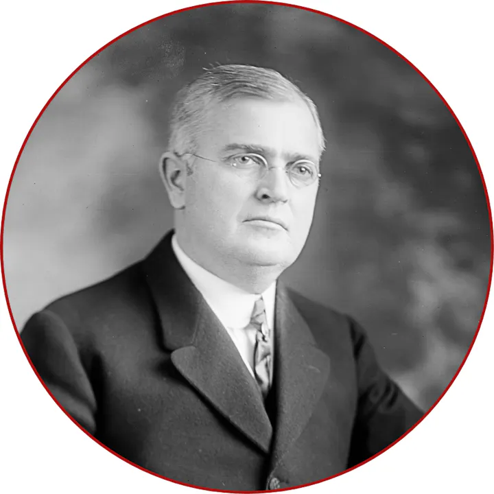 A black and white portrait of a serious-faced man in a suit and glasses, with a circular frame.