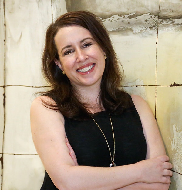 A woman smiling, arms crossed, wearing a black top and necklace, against a tiled wall background.