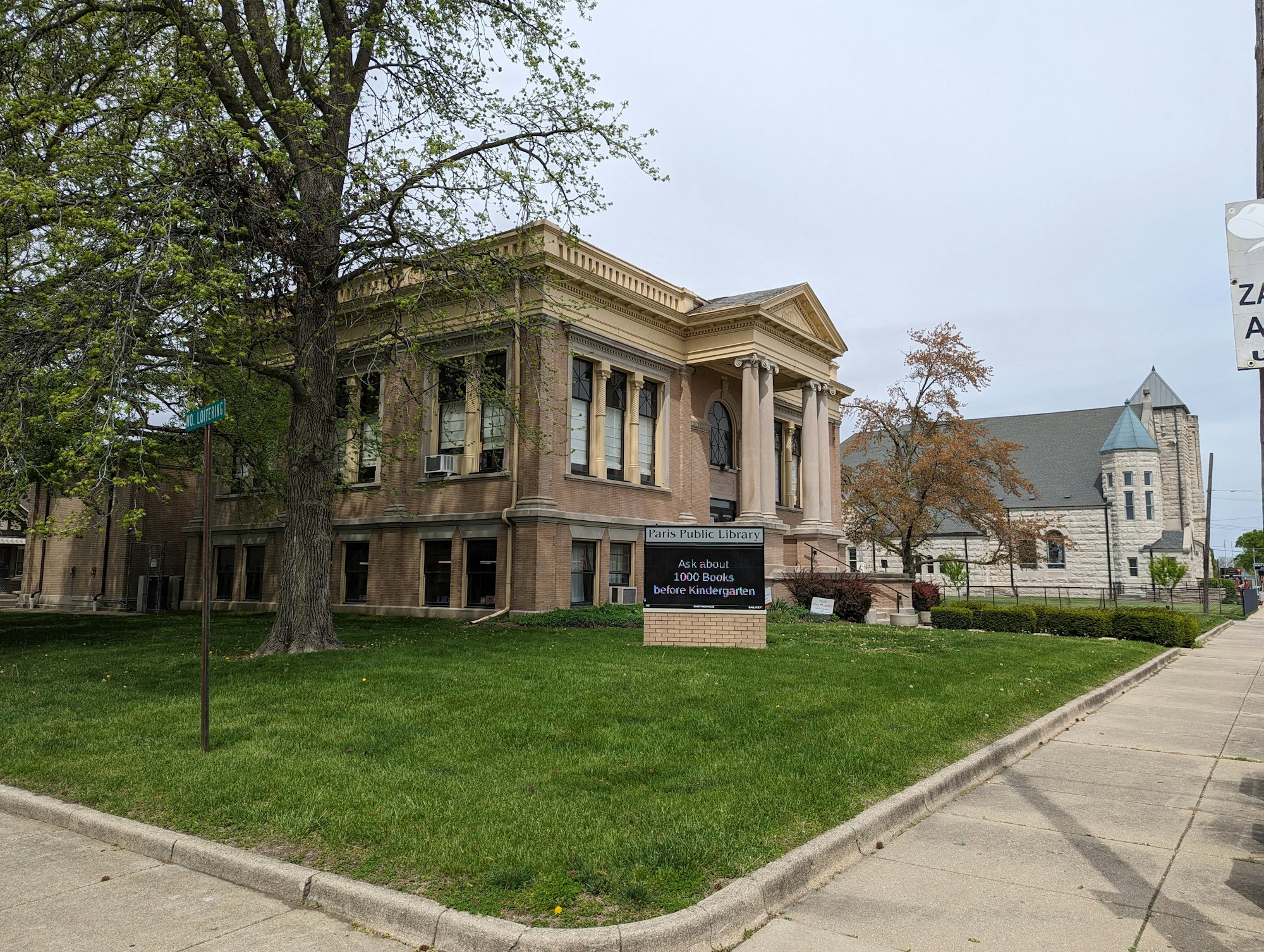 A historic library building with a grassy lawn and a sign promoting a reading program. Nearby, there's a large stone church.