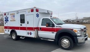 A white ambulance with red stripes and emergency symbols, parked on a paved surface.