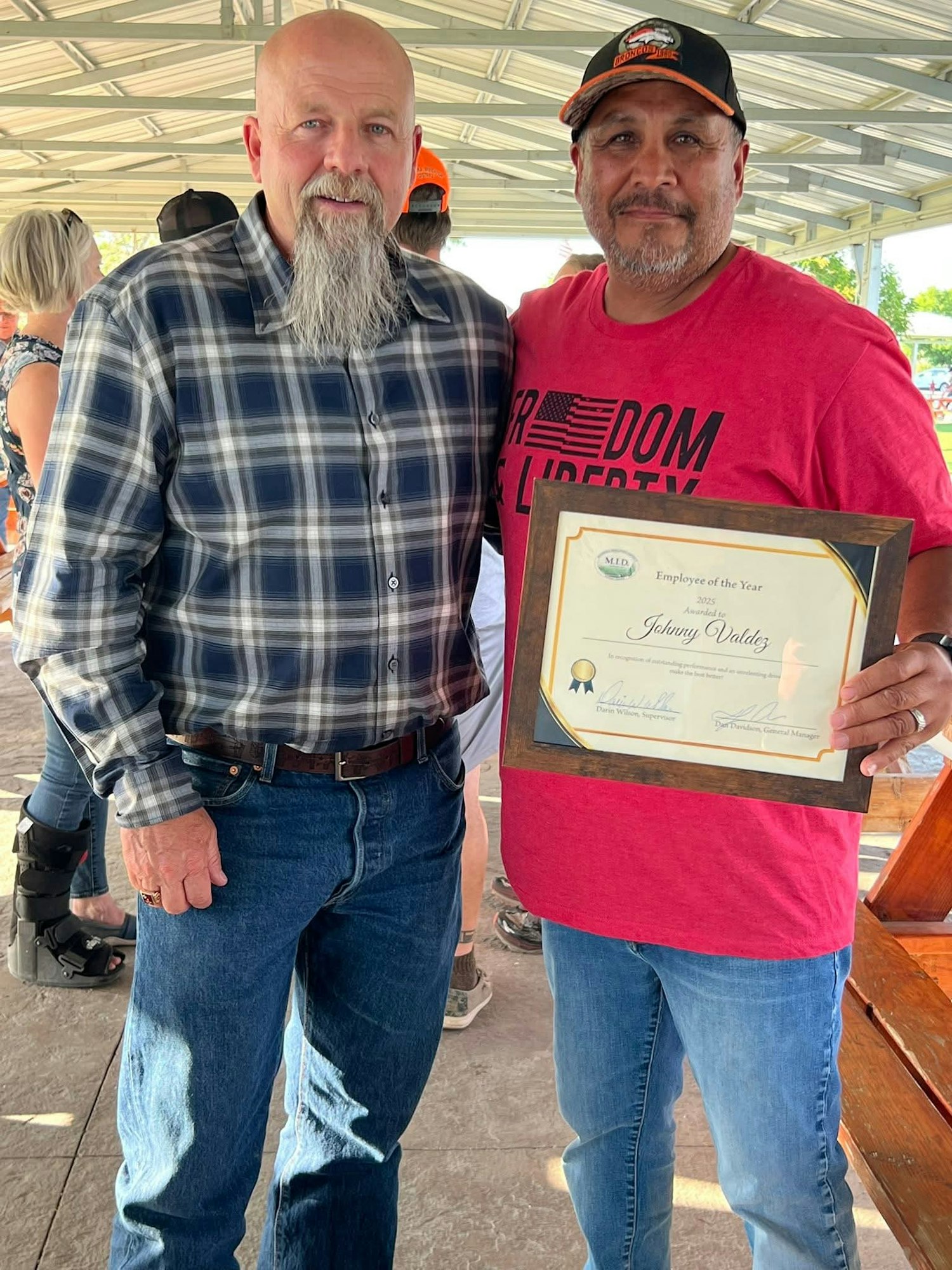 Two men smiling, one holding an "Employee of the Year" certificate. They're standing under a pavilion.