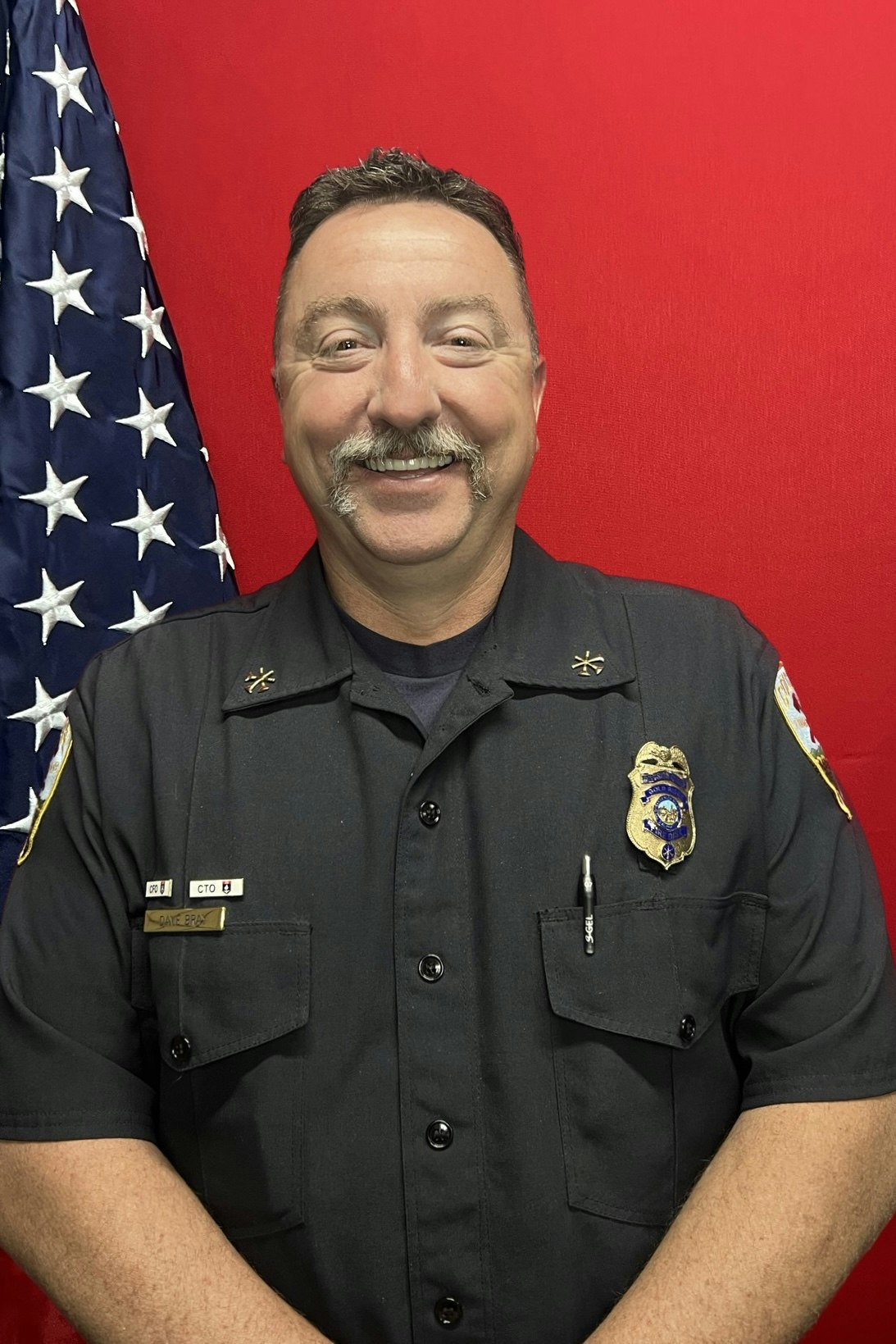 A smiling person in a uniform with badges and an American flag.