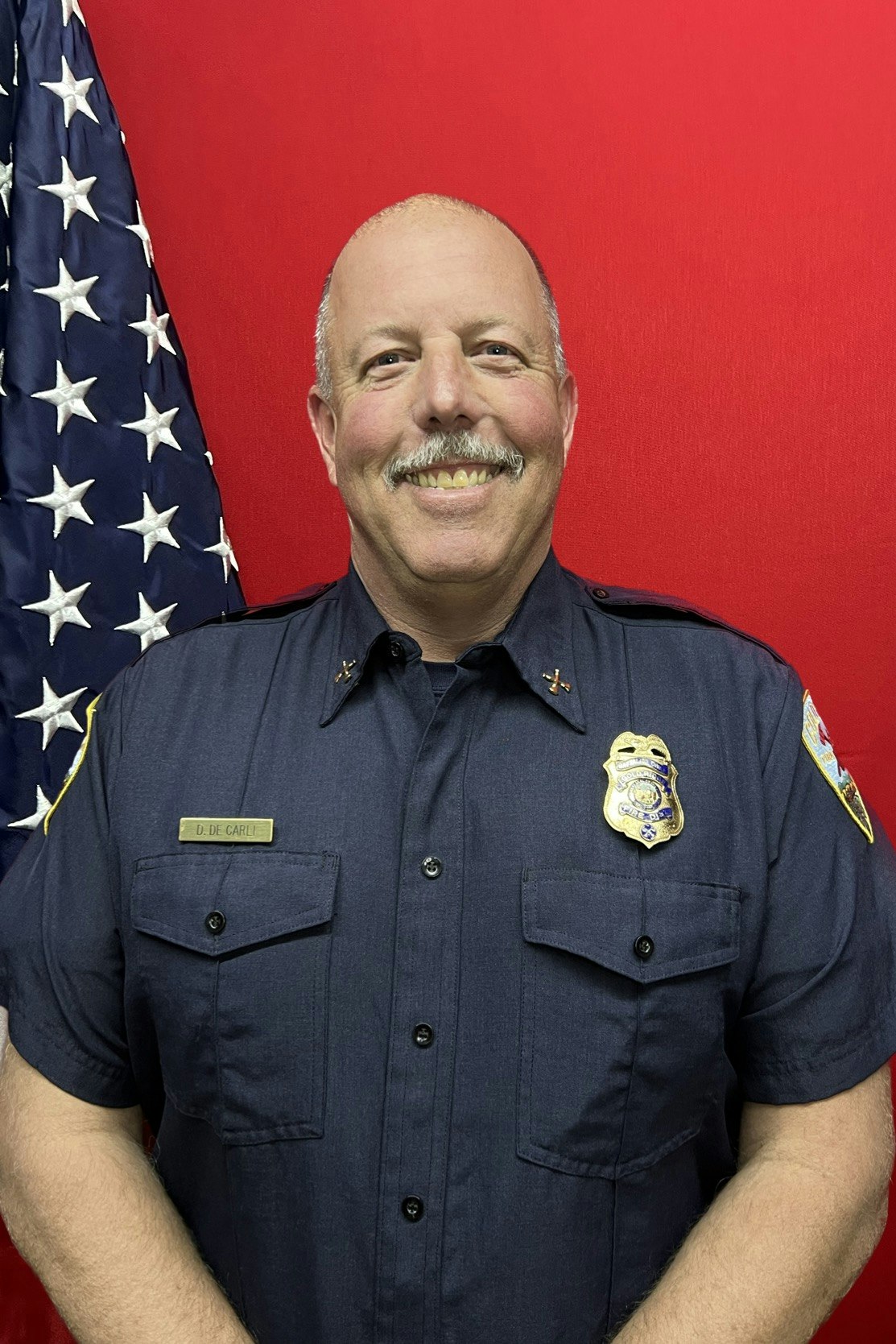 A smiling person in a police uniform with a badge and an American flag.